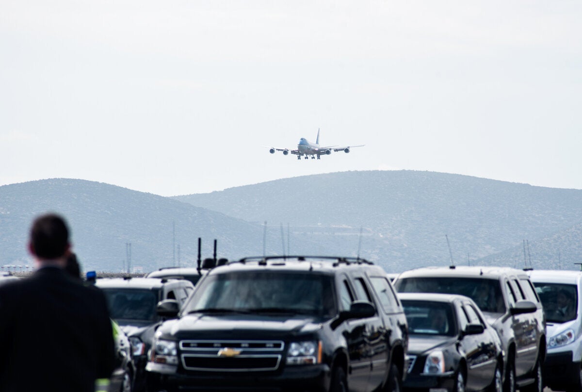 Σειρά αυτοκινήτων, βαν, και ασθενόφορων περιμένουν την ώρα που το Air Force One διακρίνεται στον ορίζοντα